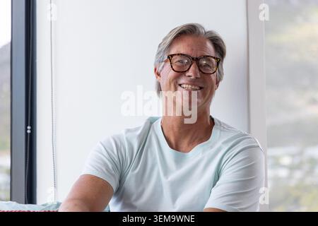 Senior man sitting by large window, smiling and wearing tortoiseshell eyeglasses, pale mint T-shirt Stock Photo