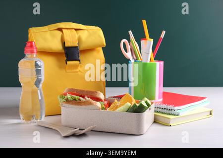 Lunch box with tasty food, cutlery, water, backpack and stationery on white wooden table near chalkboard Stock Photo