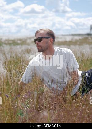 Handsome man in sunglasses relaxes on green meadow with fluffy white feather grass. Male person resting on ground and looking away, scenic wild steppe Stock Photo