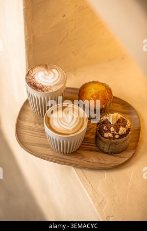 A wooden serving tray holding two hot espresso drinks with heart latte art in ribbed white cups alongside two different freshly baked muffins. Stock Photo