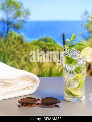 Mojito with mint, lime and ice next to a white towel and sunglasses on a terrace table overlooking a blurred Mediterranean coastal landscape. Stock Photo