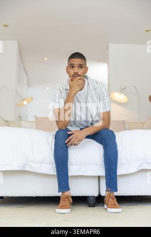 Male sitting on bed edge, contemplating with hand on chin in modern bedroom with lamps Stock Photo