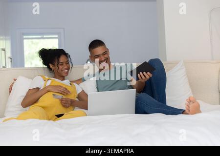 Couple reclining on white bed at home, woman in yellow overalls holding card, man using tablet Stock Photo