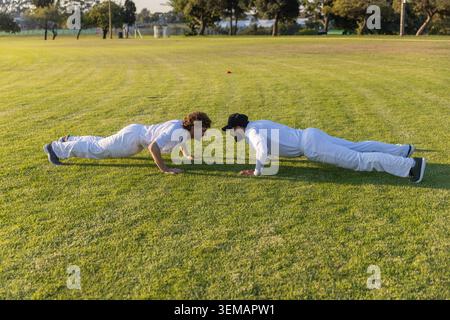 Two male athletes doing push-ups facing each other on field, white uniforms, black cap, orange cone Stock Photo