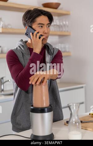Asian man holding smartphone to ear while pressing blender on kitchen counter with milk carafe Stock Photo