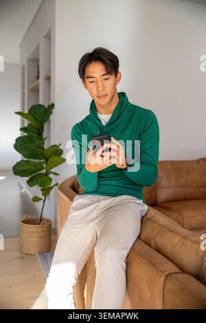 Asian man sitting on leather sofa arm in modern living room, scrolling black smartphone near plant Stock Photo