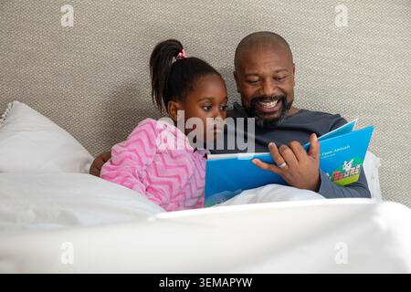 African American father and daughter in pink pajamas reading blue book on white bedding, smiling Stock Photo