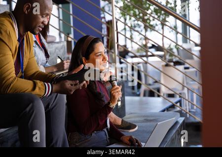 Diverse coworkers sitting on tiered steps, woman speaking into mic with laptop, man with tablet Stock Photo