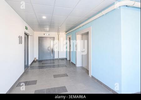 well-lit hallway features two elevators, various doors, and patterned floor tiles. One wall is pale blue, the others are white. Ceiling is textured. Stock Photo