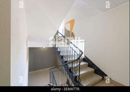 interior apartment public place, view of stairwell with concrete steps, dark railing, and white walls. An orange abstract design is on one wall. Stock Photo