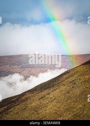 Vibrant rainbow stretches across a vast mountain vista. Fluffy white clouds float below the horizon, obscuring part of a distant rocky terrain. Stock Photo
