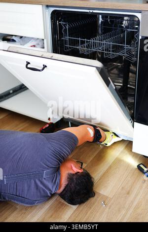 Repair worker fixing an integrated dishwasher cabinet panel with power tools during kitchen renovation and household appliance installation. Stock Photo