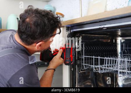 Professional technician installing a built-in dishwasher in a modern kitchen using a cordless drill during home renovation and appliance setup. Stock Photo