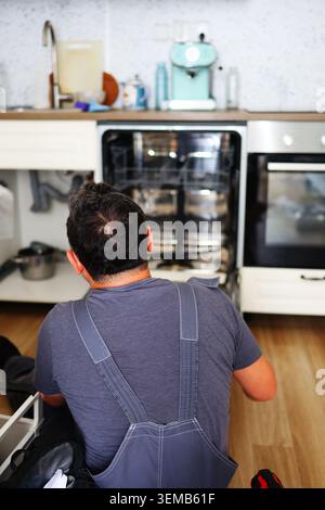 Maintenance technician working on dishwasher fitting in a modern home kitchen, highlighting repair services, renovation and construction work. Stock Photo