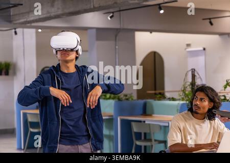 Diverse male coworkers demonstrating with white VR headset, navy hoodie, laptop in coworking space Stock Photo