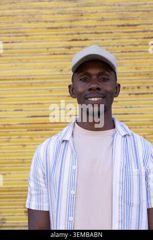 African American man standing by yellow roll-up shutter on street, wearing beige cap, striped shirt Stock Photo
