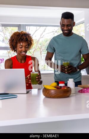 Diverse couple sipping green smoothies using laptop at kitchen counter in red sleeveless teal tee Stock Photo