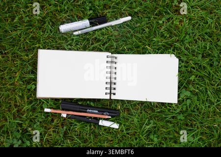 Top view of open blank spiral notebooks on green grass background, surrounded by sketching markers, drawing pencils and artist supplies. Stock Photo