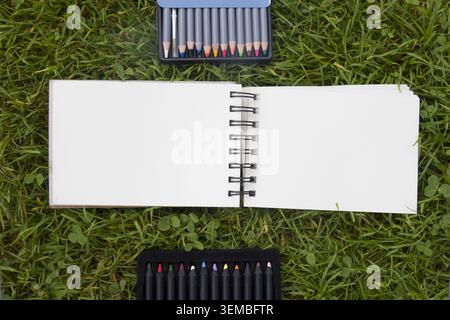 Top view of open blank spiral notebooks on green grass background, surrounded by sketching markers, drawing pencils and artist supplies. Stock Photo