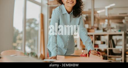 Smiling mature Hispanic female teacher leaning on a wooden desk in a bright classroom Stock Photo