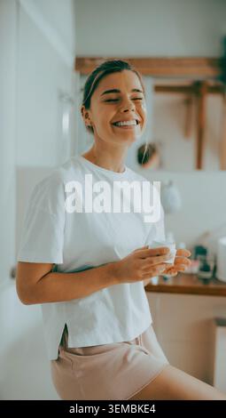 Happy young adult Caucasian woman smiling and holding a moisturizer jar in a cozy bathroom Stock Photo