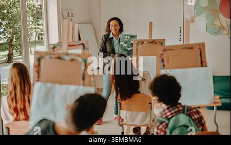 Happy Hispanic mid adult female teacher showing a painting to school children in a bright classroom Stock Photo