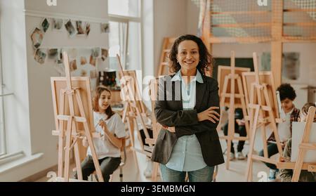 Smiling middle aged Hispanic art teacher stands with arms crossed in a bright school art studio Stock Photo