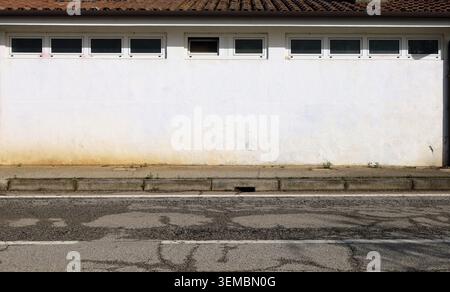 One floor building with white wall and row of small windows in the upper side. Earthenware tiles rooftop. Sidewalk and road in front. Backgrond Stock Photo