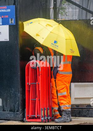 Two contractors stand close together, sheltering beneath a large, branded Aviva umbrella outside the demolition site of the St Helen's skyscraper—historically known as the Aviva Tower—on a rainy day in the City of London, UK Stock Photo