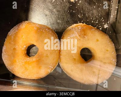 Two sugar glazed donuts sit on a polished metal shelf inside a display case, showing golden crust, sugary specks, and round holes at the centers Stock Photo
