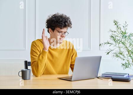 Young man in glasses using laptop for video call, gesturing with hands in confusion or explanation at home office. Curly-haired male freelancer or stu Stock Photo