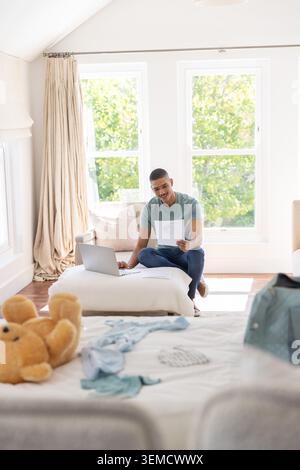 Man sitting cross-legged on ottoman, holding paper and using open laptop in bedroom with teddy bear Stock Photo