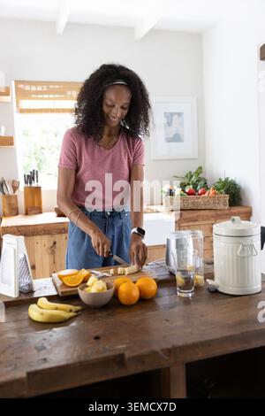 African American woman chopping fruit on wooden cutting board in white kitchen with blender pitcher Stock Photo