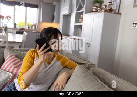 Asian woman holding smartphone, sitting on light-gray couch, turning head right during call at home Stock Photo