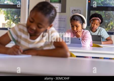 Diverse female children sitting in classroom using tablets, wearing headphones, writing with pencil Stock Photo