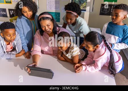 Teacher with diverse students wearing pink blouse gathering around tablet in folio at school table Stock Photo