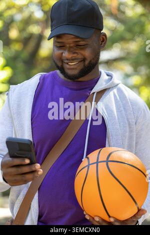 African American man holding orange basketball and checking smartphone in sunny park with bag strap Stock Photo