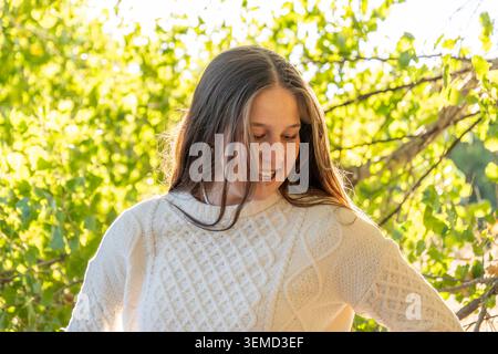 A young woman stands in a sunlit outdoor setting among green foliage, wearing a white knitted sweater with a cable pattern. She smiles softly, with lo Stock Photo