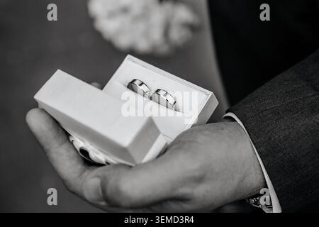 A black and white close-up shot focusing on a groom's hand holding an open gift box containing a pair of classic wedding bands. Stock Photo
