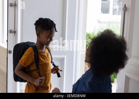 African American mother kneeling in denim jacket, smiling at son holding black backpack in foyer Stock Photo