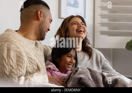Diverse family with child sitting on sofa at home, wrapping in grey blanket, wearing cozy sweaters Stock Photo