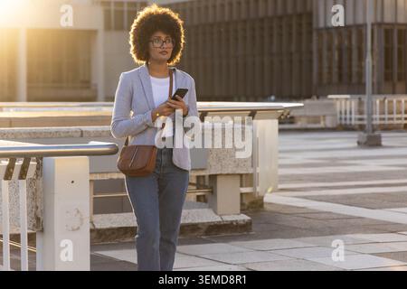African American woman in her 20s walking on rooftop, wearing blazer, holding smartphone and purse. Terrace, urban, sunlit, goldenhour, crossbody, dev Stock Photo