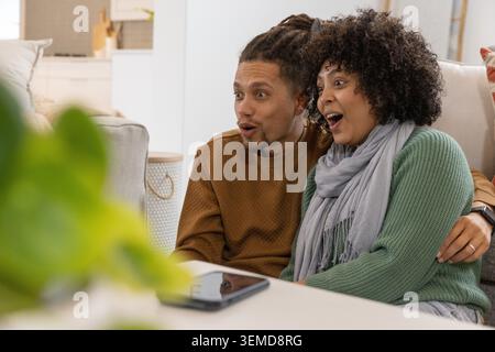African American couple sitting on couch in living room, wearing green sweater, viewing tablet. Sofa, plant, cozy, candid, watch, cushions, basket Stock Photo