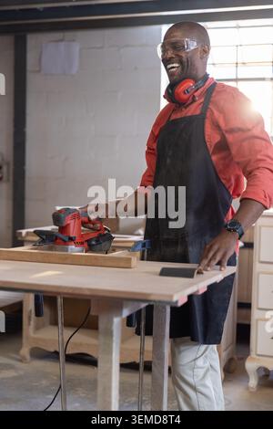 Mature African American man sanding wood plank at shop wearing goggles and using sander, copy space Stock Photo