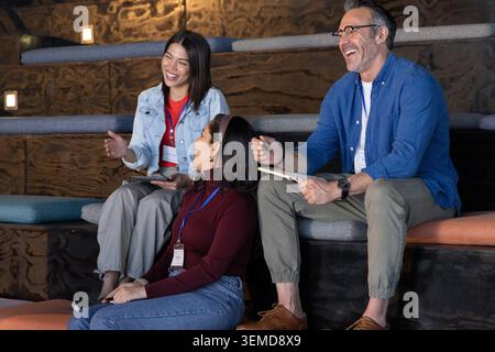 Diverse colleagues laughing, talking on tiered cushions in workshop, holding badges and notebook Stock Photo