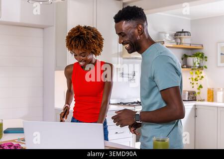 African American couple laughing and using open laptop on kitchen island with pink measuring tape Stock Photo