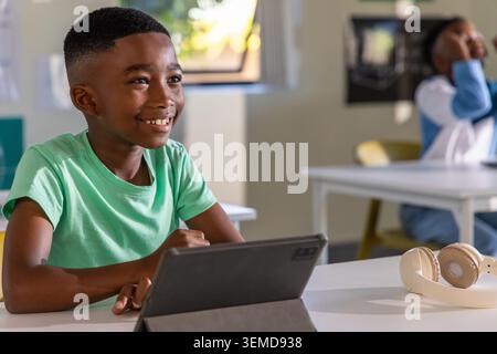 African American classmates sitting, boy using tablet at front left in class with beige headphones Stock Photo