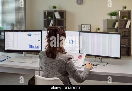 Businesswoman analyzing data on multiple monitors in modern office, charts displayed Stock Photo
