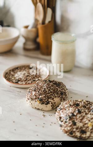 Close-up of homemade sourdough bread loaves rising on a floured countertop, topped with mixed seeds, next to ingredients in a cozy kitchen setting wit Stock Photo