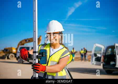 Smiling female construction worker wearing a white helmet and reflective vest uses a measuring device at a jobsite, highlighting safety, teamwork, and Stock Photo